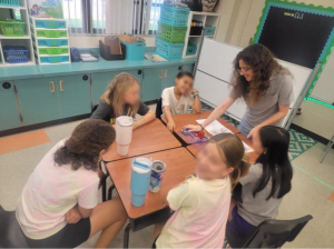 A teacher interacts with five students seated around a table covered with educational materials in a classroom. Shelves and cabinets in the background hold various supplies.